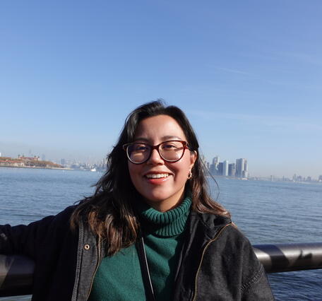 Young woman looking at camera wearing green shirt and black jacket with water in background