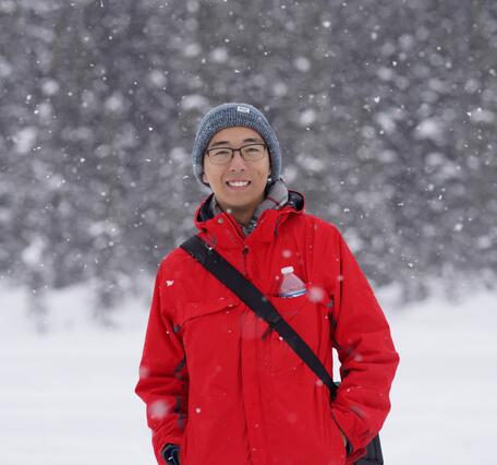 Zehao Xue stands in front of a snowy forest wearing a grey beanie and red jacket.