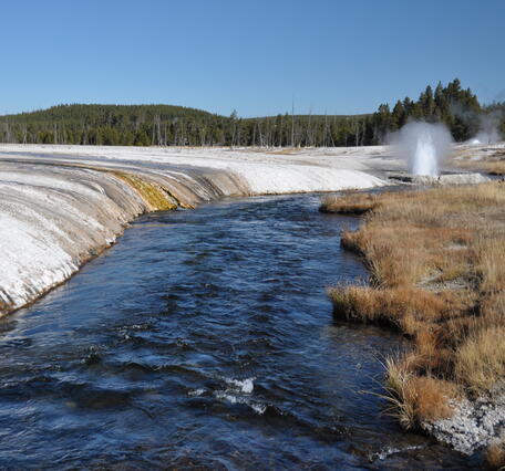 A stream winds pass a barren, white landscape on one bank and brown grass on the other; small geyser in the distance