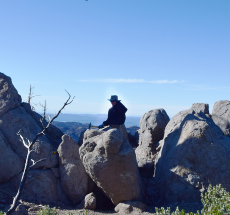 A scientist in a broad-brimmed hat sits on a large gray boulder of volcanic rock, surrounded by similar boulders. They are holding a rock hammer against the rock in preparation to break off a sample. In the distance, several hazy mountain ridges can be seen. 