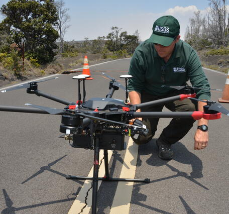 a man in dark green USGS gear kneels next to a large black drone in the middle of a road, cones and trees in the background