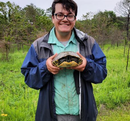 Dylan Wichman photo. Man with dark hair and glasses wearing blue jacket and green shirt standing outdoors holding a turtle