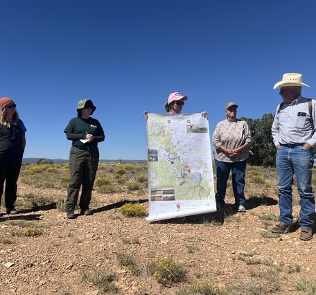 Five ranchers and researchers stand outdoors in a field looking at a large map, discussing land management