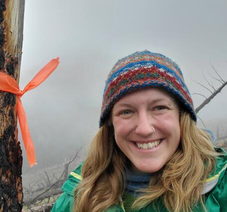 photo of person with long blonde hair in green jacket and multi-colored beanie, tree and clouds behind them