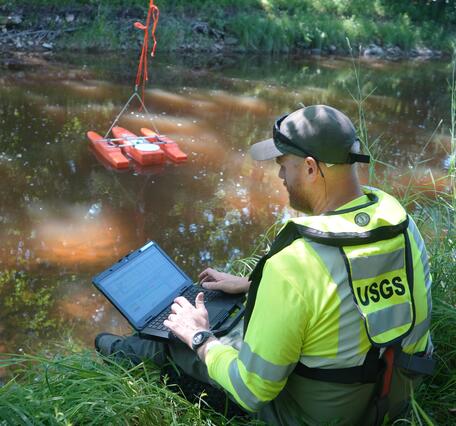 Hydrologic Technician sitting next to stream monitors streamflow data transmitted from an acoustic Doppler current profiler