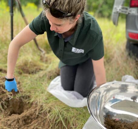 blonde lady in green USGS shirt wearing latex gloves uses a trowel to dig in grassy field
