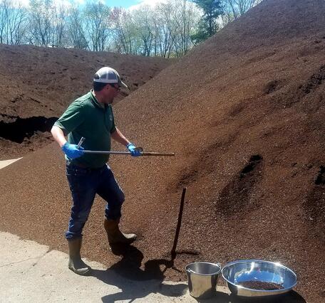 A man in green shirt and white baseball cap digs with shovel into a towering pile of dark brown dirt looking material