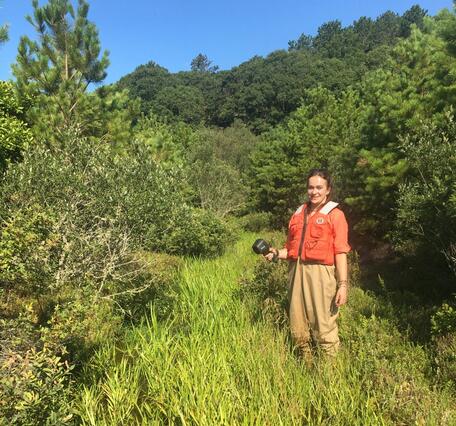 A lady in waders and orange life vest stands in a grassy bog surrounded by trees on a blue sky day