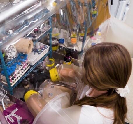 Dark blonde female in lab gear reaches into a chamber surrounded by glass bottles and other lab equipment
