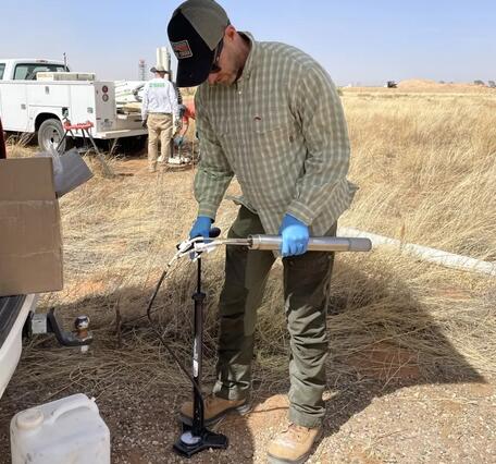 Male in baseball gap, glasses and latex gloved hands standing next to truck in a dry grassy field prepares an instrument