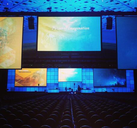 Backlit conference backdrop and stage and a row of seats in the foreground in a low-lit ballroom