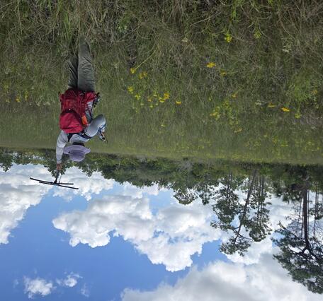 a person with red backpack and purple hat holds up a tracking device in a field with yellow flowers, trees in background