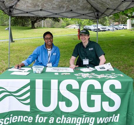 Two USGS employees smile while sitting behind a USGS tablecloth covered table under a pop up tent