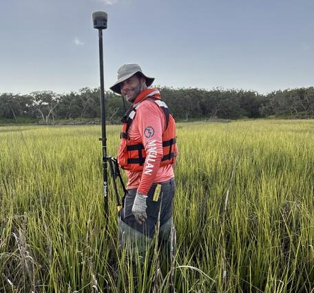 USGS scientist, Nicholas Enwright, holds survey equipment on a tall pole as he stands in a grassy marsh. He is wearing a brown bucket hat, a red long-sleeved shirt, and a orange life jacket. Trees can be seen in the distance under a blue sky with scattered clouds.