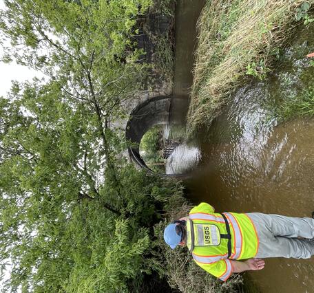 A USGS technician in a bright green safety jacket takes a photo of a flooded river