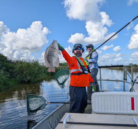 two people in life vests stand in a boat - one holds a fish while the other holds a dipnet