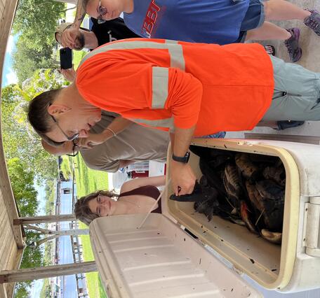 A group of people stand around a cooler filled with nonnative fish as a man in an orange shirt gives identification guidance