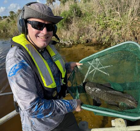 a man holding a fish in a net stands with water and vegetation seen in the background