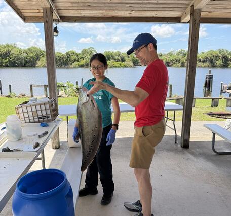 two people stand near water as they prepare to preserve a large clown knifefish in a large blue jug that sits nearby