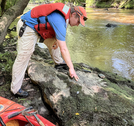 A usgs geologist in a bright red PFD examines a large boulder along a creek. 