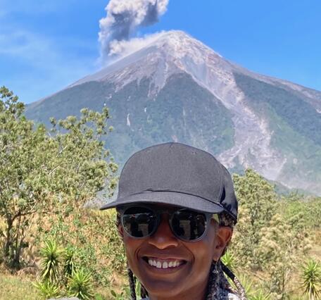 woman with black baseball hat with a volcano erupting ash in the far background.