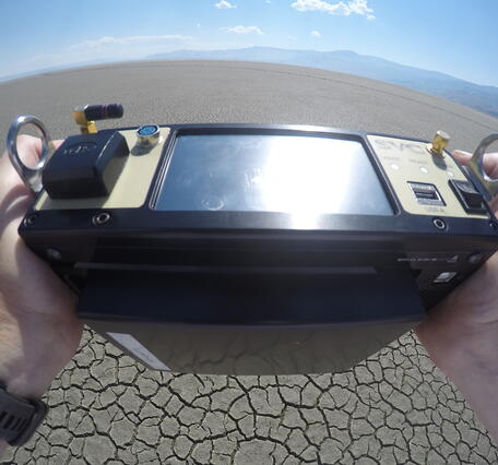 Person holds a rectangular device in both hands pointing it at the dry desert landscape.