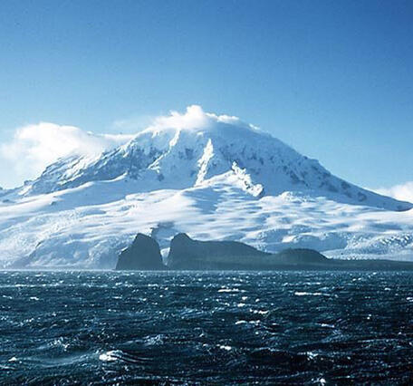 Ice-covered mountain  under blue sky with choppy dark blue ocean water in the foreground