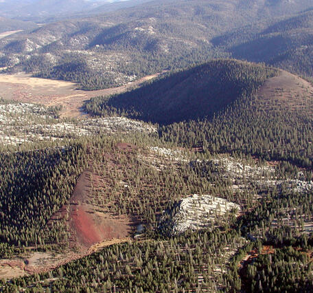 A small forested scoria cone with reddish rocks drapes over bright white outcrops of Sierran granite. The cone sits in a valley amid low rolling hills backed by higher granite peaks. 