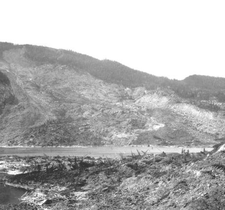 Black and white photo of a hillside that has been devastated by a landslide, with rock debris in the foreground and a lake