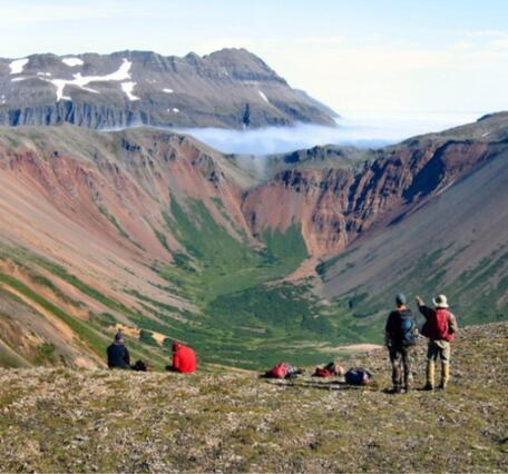 Two people standing by a ledge facing and gesturing away towards mountains in the distance; packs and two people on ground