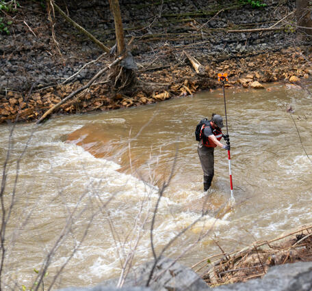 A man stands in a stream with surveying equipment.