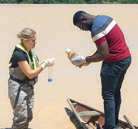 USGS scientist standing in shallow brown river training Ghanaian scientist standing on wood canoe on water sample collection