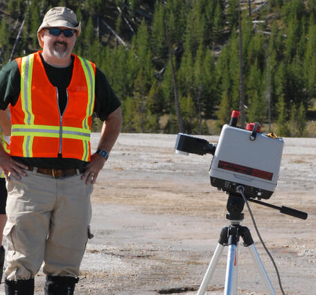 A man wearing a reflective vest and standing on barren ground stands next to a tripod bearing equipment in front of a forest