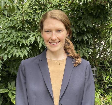 Woman in grey jacket and tan shirt standing in front of green plants.