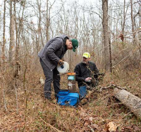 In a forest, one person measures a groundwater well while the other one inputs data into a laptop.