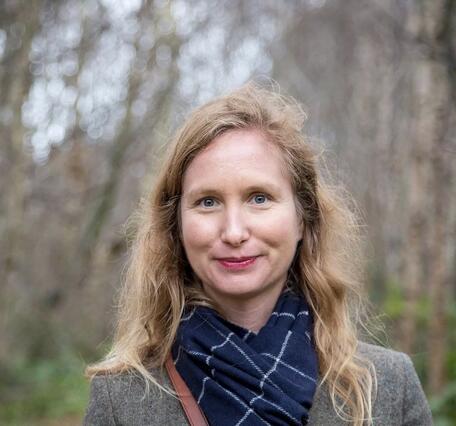 A woman with blonde hair wearing a jacket and scarf with trees in the background.