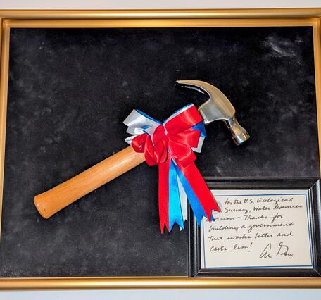 Hammer with a red, white, and blue bow mounted on black felt canvas with a golden frame