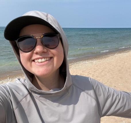 Person wearing sunglasses and hooded shirt on beach, smiling for camera