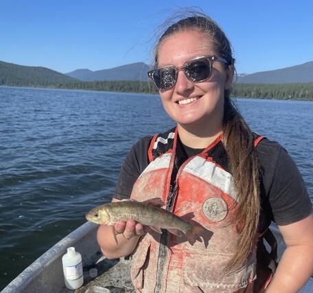 Girl with sunglasses and life jacket holding a fish on a boat. Water, trees, and mountains in the background.
