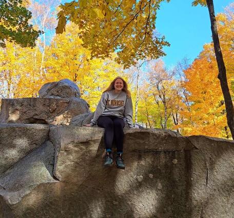 Woman sitting atop a rock formation with trees in fall colors in the background.