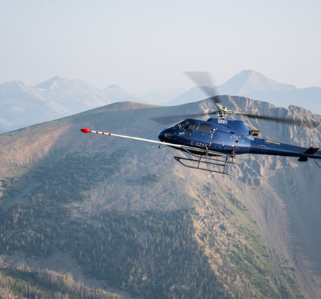 A blue helicopter flies in front of mountain with a long, white boom arm sticking out the front of the aircraft with red tip