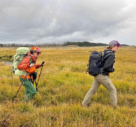 Two people wearing raincoats and hiking across a grassy meadow under cloudy skies