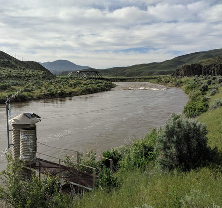 USGS gage house on the bank of the Humboldt River, with green hills surrounding the site and a cloudy sky overhead.