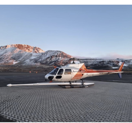 Red and white helicopter with boom extended, on surface in front of mountains.