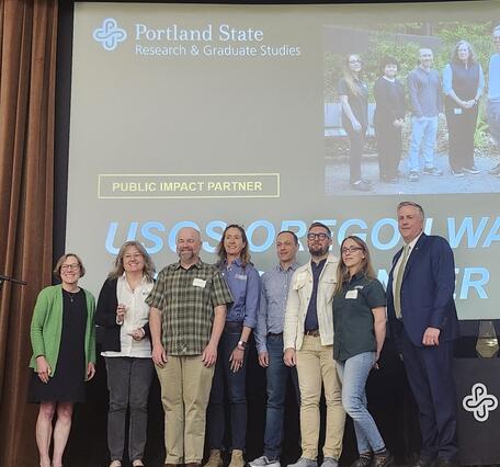 8 people stand on a stage in front of a projection screen with award info on it