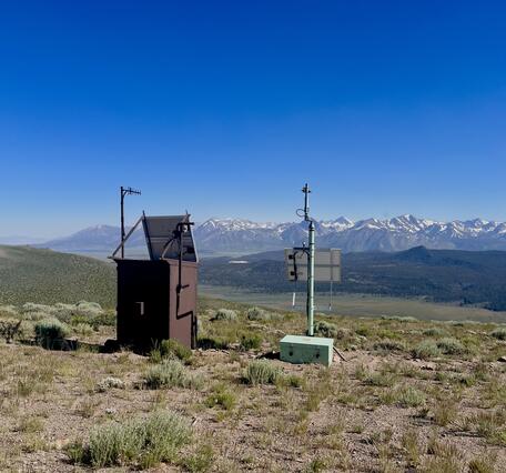 In this photo, a large brown metal box and a small aqua-colored box are both topped by antenna poles and solar panels. They sit side-by-side on a sagebrush-covered hill overlooking a broad valley and a line of jagged, snow-capped mountains.