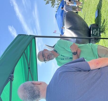 USGS scientist talking to a man next to a table, under a tent, outside by the water.