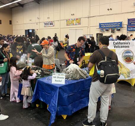 This photo shows a science festival booth surrounded by children and their families. A model volcano sits on a blue tarp behind a "Mt Shasta Rocks!" sign. Behind the table, a scientist in an orange USGS cap explains volcanic rocks, while another scientist in a volcano-themed Hawaiian shirt prepares the model volcano for its next "eruption".