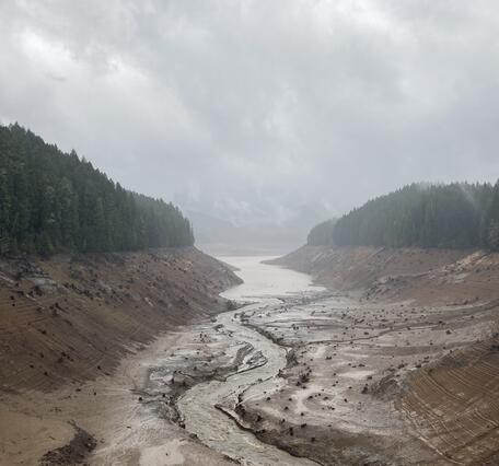 Cloudy rainy day. Pine trees top brown steep muddy banks of empty reservoir with stream running through muddy channel
