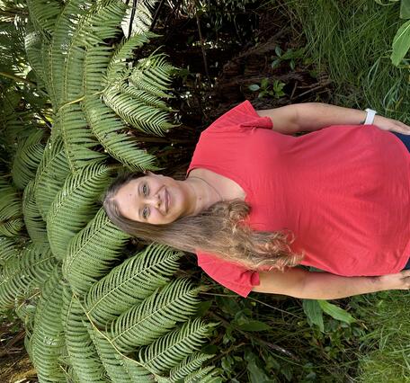 Person in orange shirt with long brown hair in front of tree ferns.